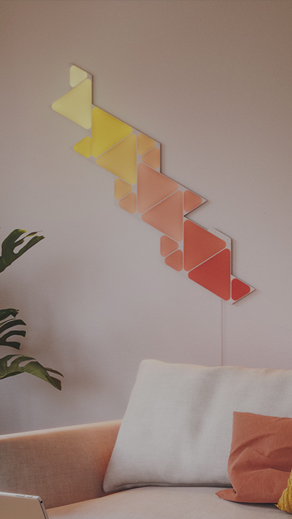 Questa è un’immagine dei pannelli Nanoleaf Shapes Triangles e Mini Triangles in un soggiorno, sulla parete sopra il divano. I pannelli luminosi smart che cambiano colore e modulari sono collegati tramite connettori e dispongono di oltre 16 milioni di colori.