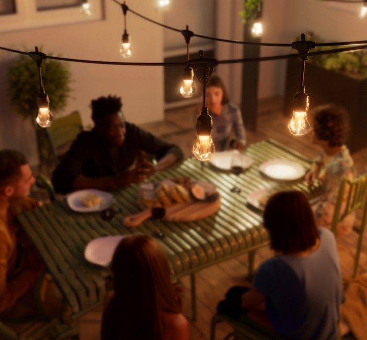 Cozy outdoor patio with six people gathered. Smart outdoor string lights casting a warm yellow glow above a table with a nearby BBQ grill and plants around. A charcuterie spread is on the table with plates set out.