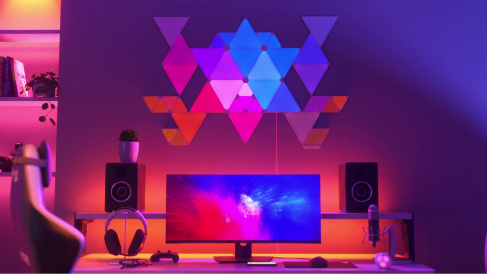 Dies ist ein Bild der Nanoleaf Shapes Triangles und Mini Triangles an der Wand über dem Schreibtisch und hinter dem Monitor einer Gaming-Battlestation. Diese RGB-Lichter verfügen über 16 Mio. Farben und sind perfekt für den Gamer daheim. Die modularen, intelligenten Light Panels werden mithilfe von Verbindungsstücken miteinander verbunden, um ein Design zu erschaffen.